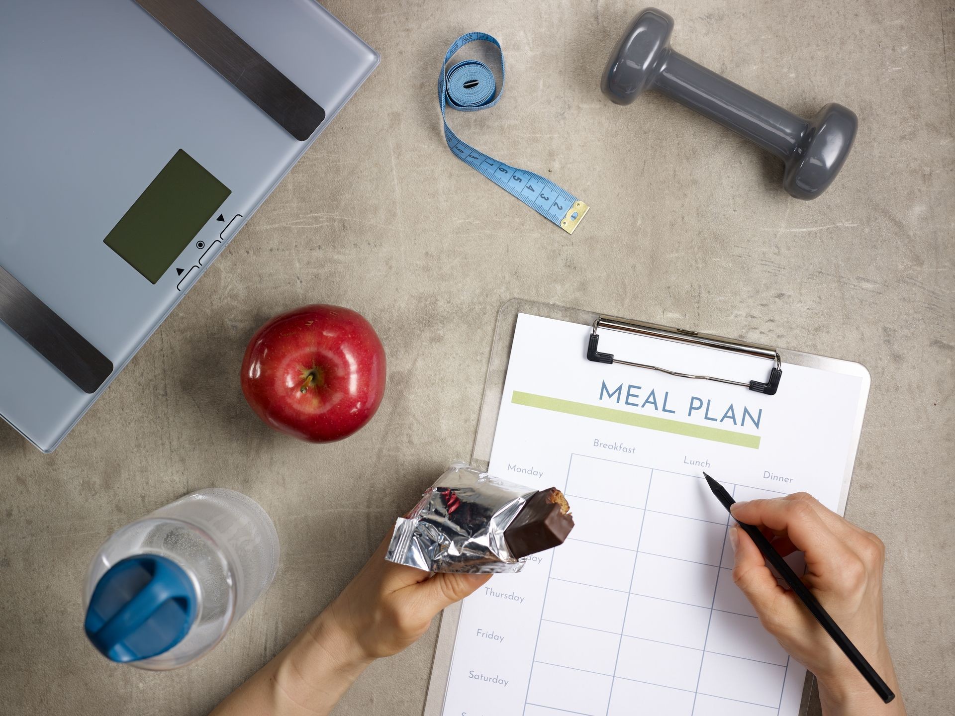 Closeup on weight scales, grey dumbbell, red apple, bottle of water, tape measure laying on the floor and female hands with bitten raw protein bar filling meal plan.